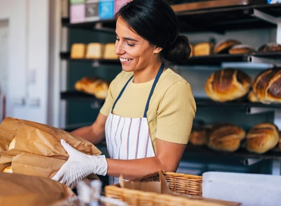 A cheerful baker in an apron packs fresh bread into paper bags in a bustling bakery. The scene conveys warmth and artisanal craftsmanship.