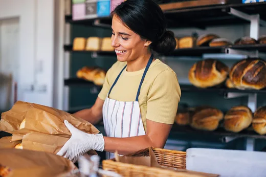 A cheerful baker in an apron packs fresh bread into paper bags in a bustling bakery. The scene conveys warmth and artisanal craftsmanship.